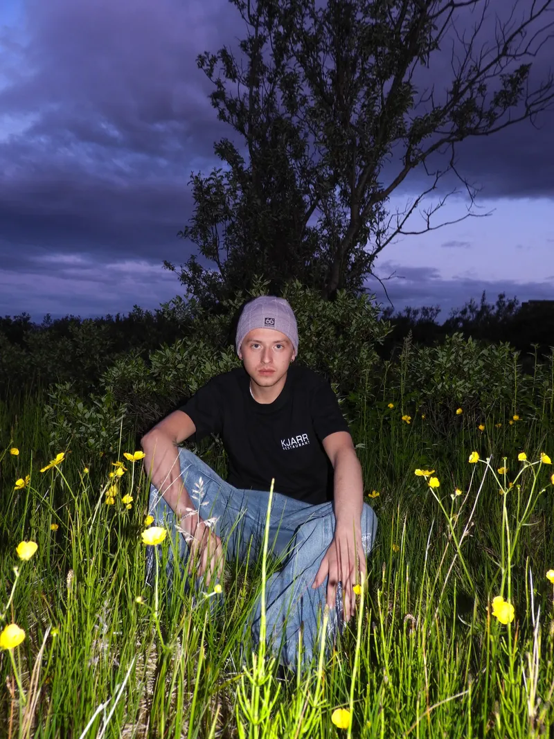 Young man in a beanie crouching in a field of buttercups at dusk