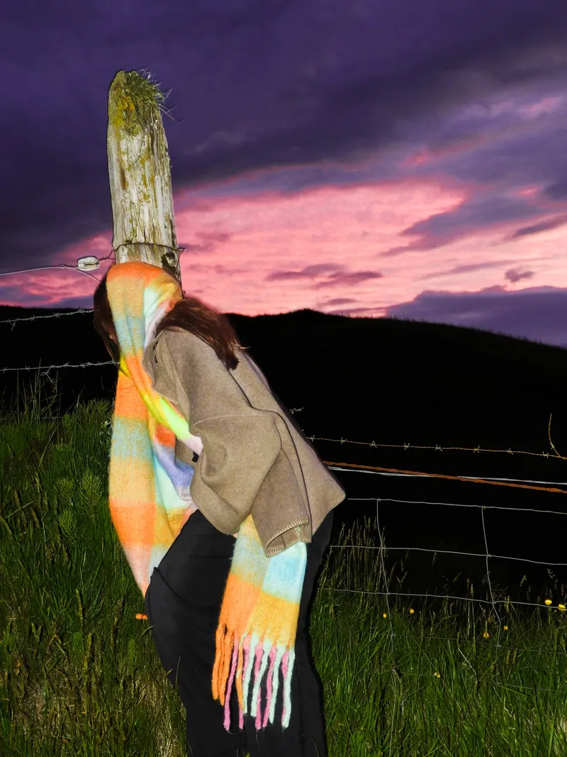 Woman in a rainbow scarf leaning on a fence post against a pink sunset