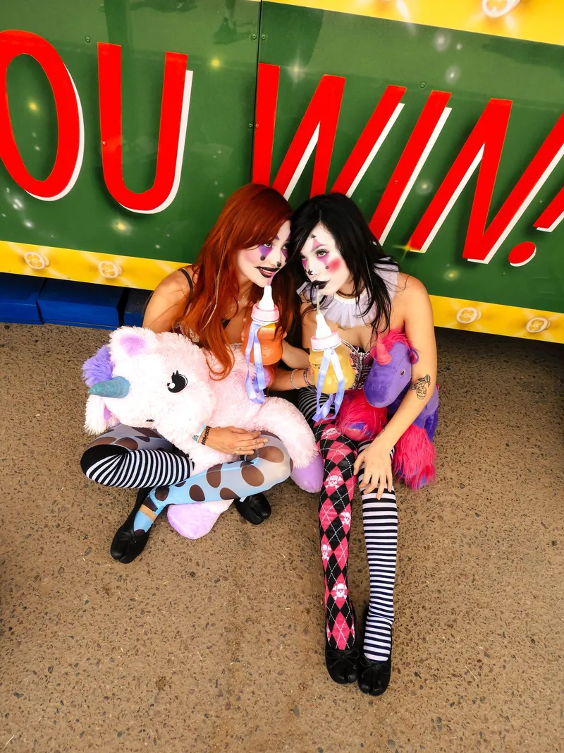 Two women in clown makeup with plush unicorns under a YOU WIN carnival sign