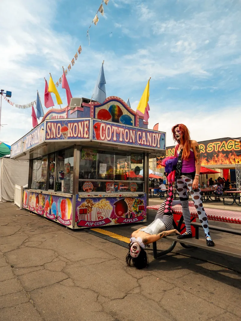 Two women in clown makeup at a carnival snack stand under a blue sky